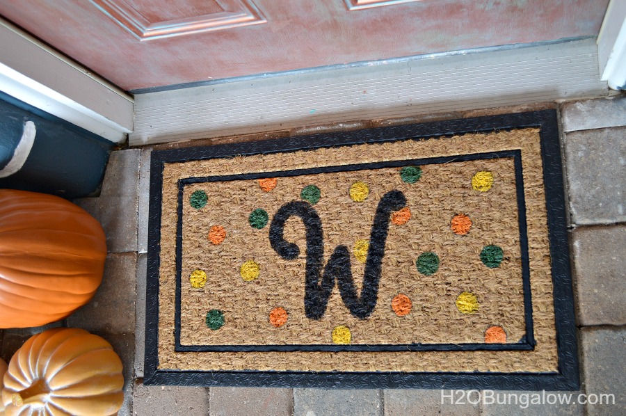 simple fall monogram doormat in front of copper door with orange pumpkins next to it.