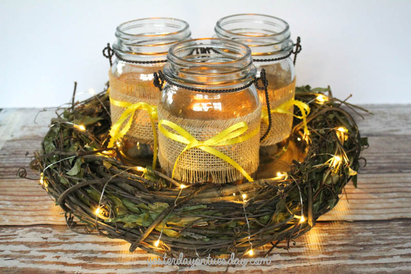 3 mason jars with burlap sleeves sitting in the middle of a wreath wrapped in mini string lights