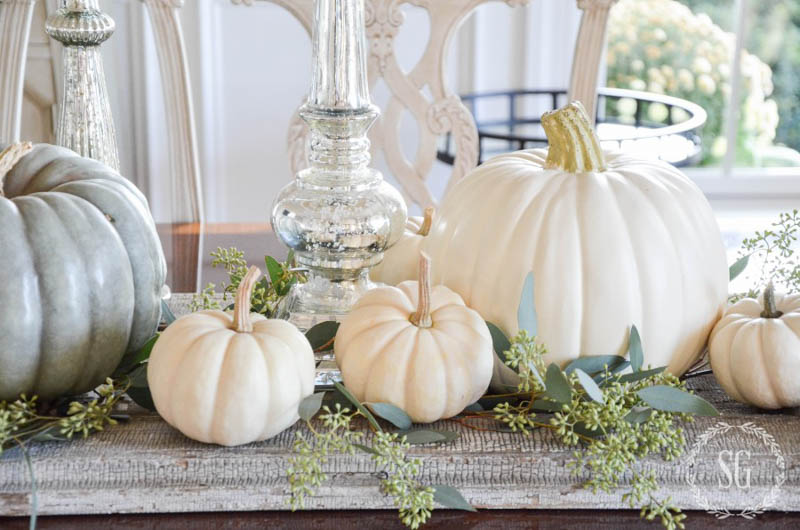 White pumpkins sitting on a table runner with greenery and clear glass candlesticks