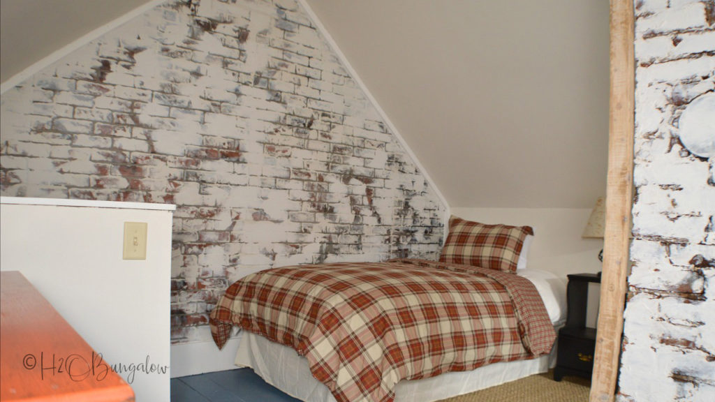 Brick wall with white german schemer with bed and red checked bedspread in front of it.