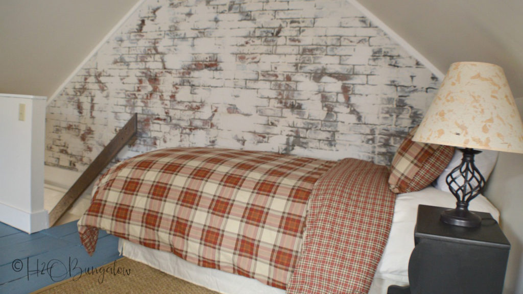 Bed with red checked bedspread, nightstand with lamp with view of stairs going down out of the loft