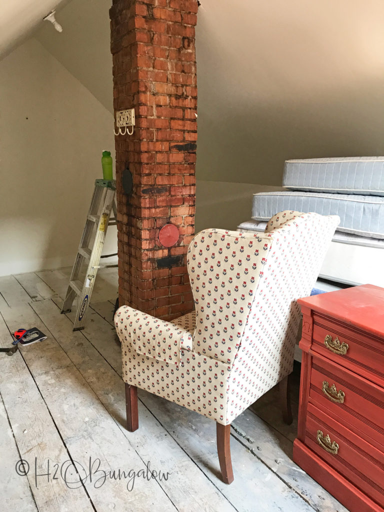 brick chimney in loft with wing backed chair sitting next to it and red dresser and mattresses
