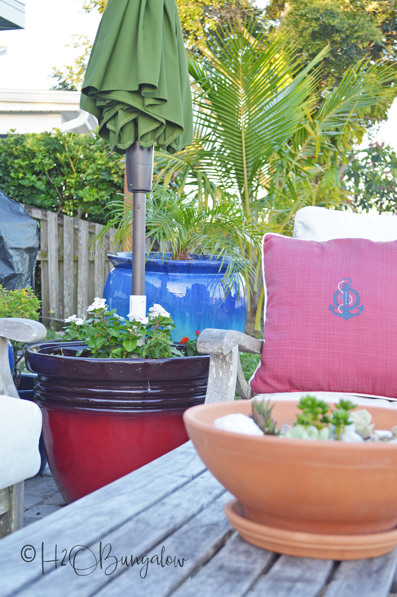 A DIY umbrella planter stand is an easy spring outdoor DIY project.  Sitting next to next to a blue planter and chair with pink pillow.