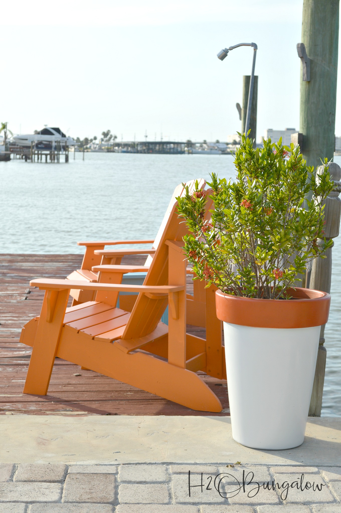 White planter with orange rim sitting next to orange Adirondack chairs on a dock by the water.