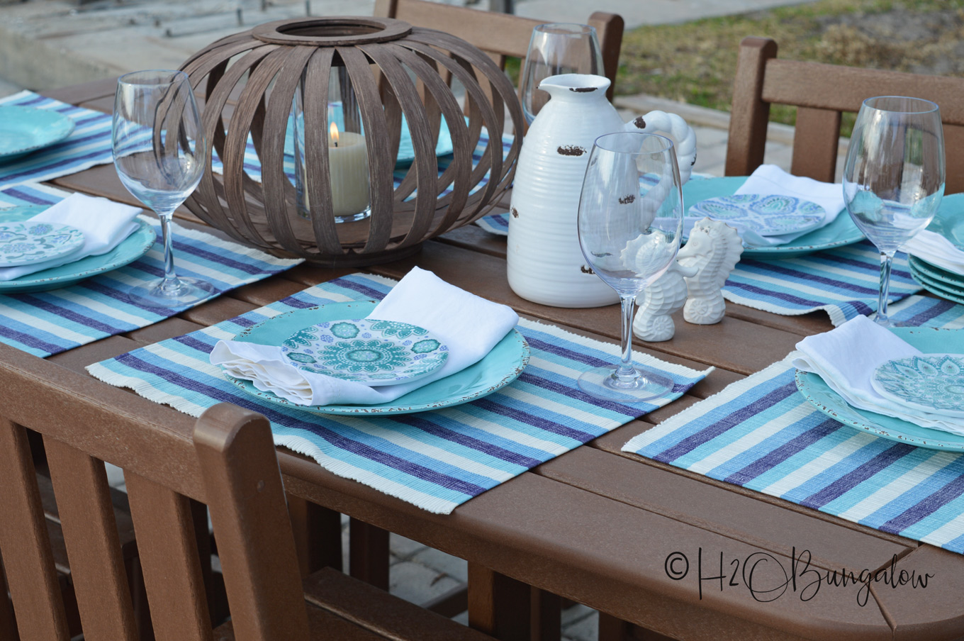 Outdoor dining table set with blue and white striped placemats and coastal colored dinnerware and wooden lantern as part of the centerpiece.