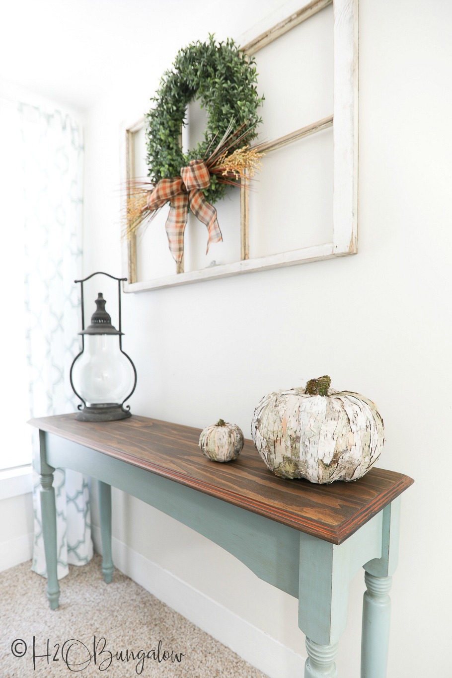 Birch bark pumpkin sitting on console table with lantern and box wreath hanging above on old window.