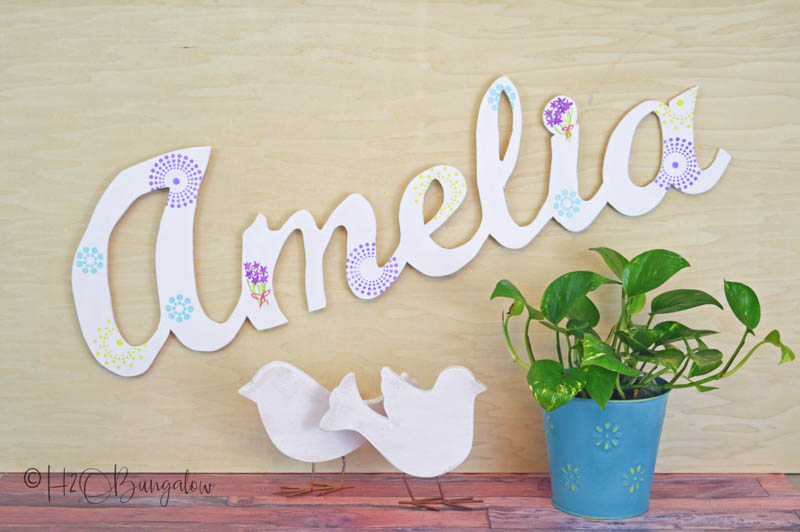 Wooden name cutout "Amelia" on the wall with wooden birds and a plant on the table below